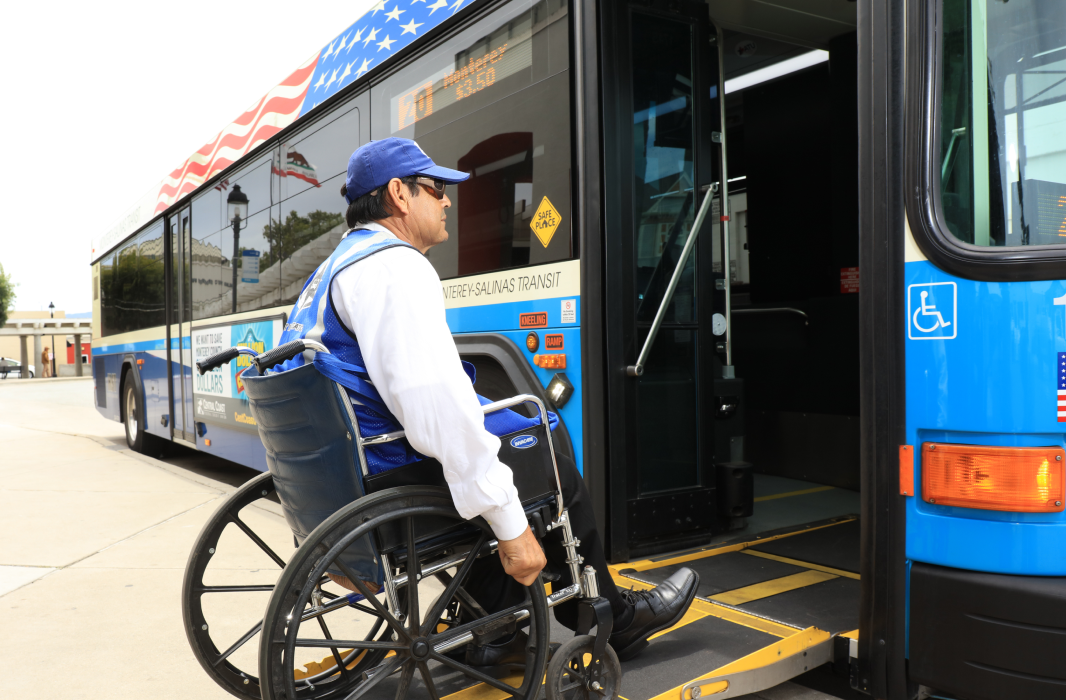 A man in a wheelchair boarding a MST bus via a wheelchair lift. The bus is blue with a waving American flag decal across the top. The bus is making a stop at a wide sidewalk.